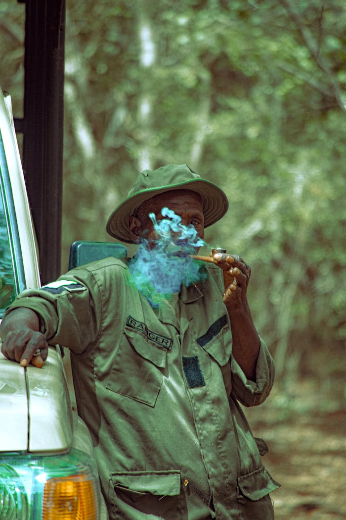 A ranger in Mainamaji, Nigeria, wearing a green uniform and hat, stands smoking in the forest.